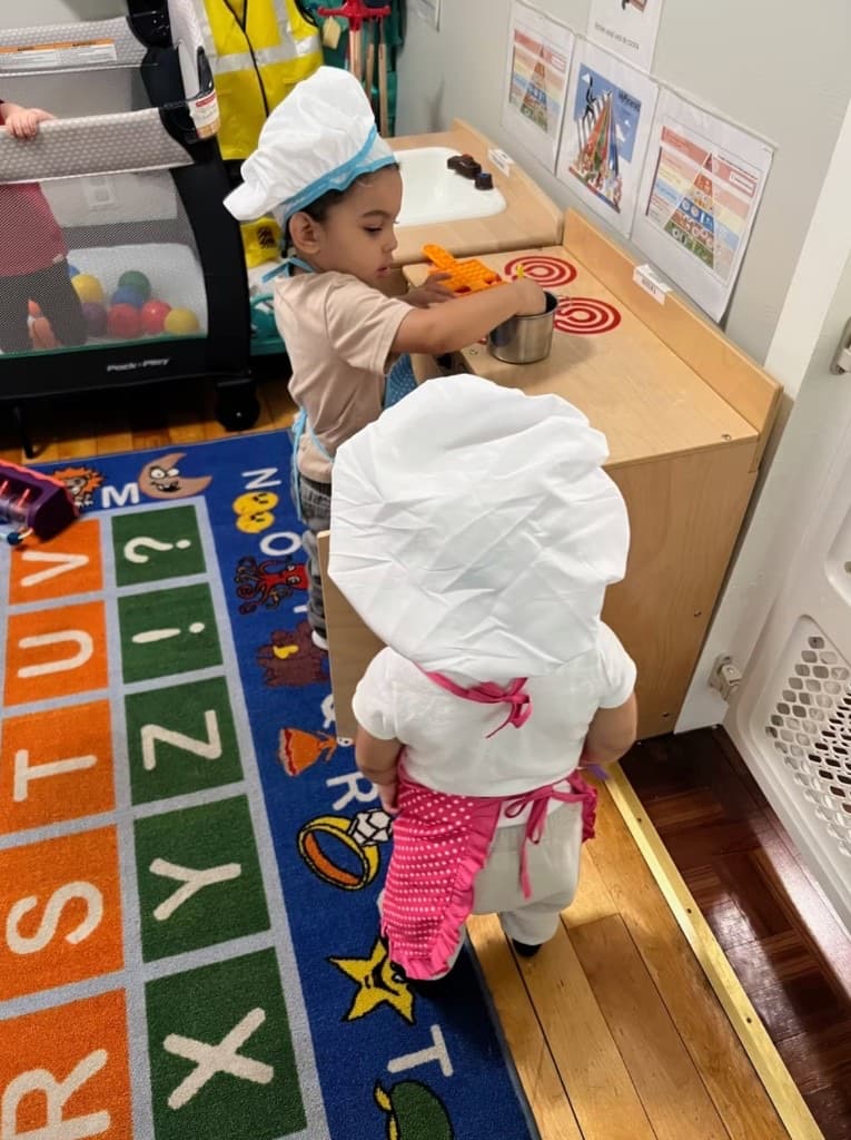 Toddlers playing in chef hats at a wooden play kitchen
